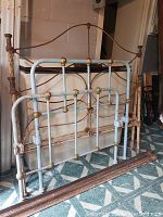 Two antique iron bed frames positioned against a wall, one blue and smaller, one rusted and larger, with a set of railings lying in front.