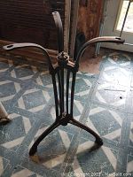 Cast iron café table base with three curved mounting arms and tripod legs, placed on a tiled floor in a room with wood panel walls and a window.