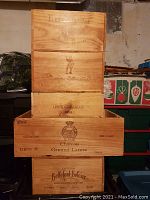 Photo showing five stacked wooden wine crates featuring printed wine labels and vintage years around 2003. Crates have different wine brand logos and text, some with crowns and decorative elements.