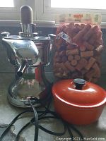 Photo showing the Healthmaster chrome juice extractor with electric cord, the orange Le Creuset enameled cast iron saucepan with lid, and a mesh bag filled with assorted wine corks on a counter.