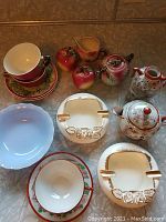 Shows 2 hand-painted apple motif cups and saucers, apple shaped salt and pepper shakers, cream and sugar set, blue Pyrex bowls, Japanese hand-painted teapot and other china pieces, and two rose ashtrays.