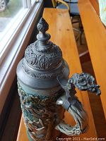 Close-up of the pewter lid of the tall stein showing intricate engravings and flame finial, with hand-painted body showing ornate scenes and floral designs.