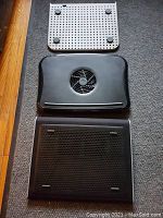 Photo showing three laptop cooling pads side by side on the floor, two black and one light-colored, two of which have fans and one without.
