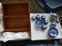 Photo showing the wooden display shelf lying open alongside the dissected tea set pieces including cups, plates, teapot, sugar bowl, and creamer.