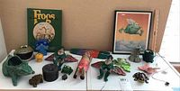 Overview photo of various frog collectibles including figurines, pottery, art prints, and decor items displayed on a table.