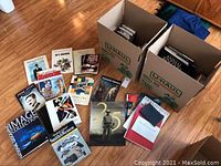 Photo showing an assortment of large hardcover books laid on floor and two partially filled boxes containing more books.