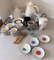 Full lot view showing all teapots, cups, mugs, small plates, and gold teaspoons arranged on table.