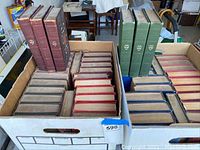Boxes containing 50 volumes of The Harvard Classics, showing a variety of binding colors: red, green, blue, brown, and maroon with some damage to edges.