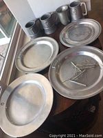 Four pewter mugs, three platters, one plate, and four plate hangers arranged on a wooden surface near window light.
