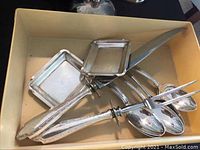 Photo of a box containing various sterling silver serving utensils including a carving fork, carving knife, and small trays with decorative handles.