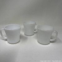 Three white opaque milk glass Fire King coffee cups with wide handles arranged on a plain surface.