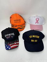 All four baseball caps arranged on a flat surface showing front designs; orange Meadowland Farmers Coop, white pink ribbon, black Bikerweek Sturgis with flag flames, navy USS Missouri BB 63.