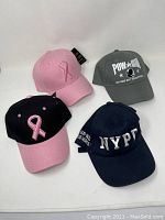 Four baseball caps shown in single image: pink ribbon solid pink, black and pink ribbon, NYPD blue cap, POW MIA grey cap