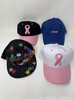 Four baseball caps arranged in a 2x2 layout. Top row includes a black cap with a pink breast cancer ribbon and a blue eBay cap. Bottom row includes a black Chicago puzzle piece autism awareness cap and a white cap with a pink breast cancer ribbon.