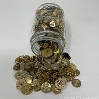 Mason jar tipped over with assorted gold-tone buttons spilled out showing different designs and shapes.