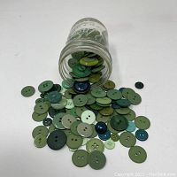Glass Ball jar filled with assorted green buttons of different sizes and shades, some spilling on a white surface.