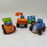 Three plastic toddler construction toy vehicles: an orange dump truck, orange excavator, and blue tractor with green trailer displayed on white background