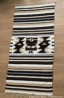 Full view of the Mexican blanket laid out on wooden floor showing striped pattern and central panel with figure and geometric shapes.
