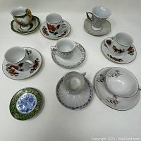 Top and angled view of eight miniature cups and saucers with various floral and decorative patterns, arranged on white surface.