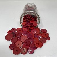 Glass jar tipped over with red buttons spilling out, showing variety of sizes and hole configurations.