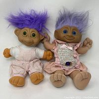 Front view of two Russ troll dolls, one male with bright purple hair and overalls, one female with light purple hair in floral pink dress.
