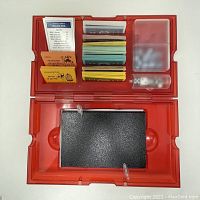 Open red plastic case showing Monopoly game board, cards, play money, tokens, dice, and instruction booklet.