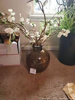 Full view of round brown glass vase with white artificial flowers and branches inside, placed on carpeted floor near window.