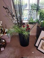 Full view of large black pot with artificial grass, broad leaves, red flowers, and metal rods with scrollwork and pinecone decoration