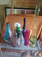 Photo showing seven assorted glass vases on a metal shelf with a wrinkled orange fabric background.