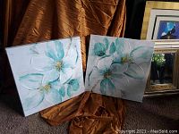 Two floral canvas paintings displayed standing against a draped brown fabric with a glimpse of framed artworks behind