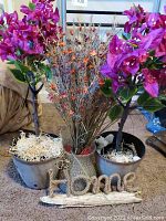 Photo showing two artificial potted plants with purple flowers, a floral arrangement with orange flowers in a vase, and a woven Home sign on the floor.