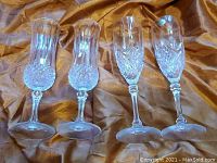 Four champagne glasses displayed on a gold-brown fabric, two with crystal cut design and two with etched glass design.