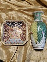 Square decorative plate with intricate multicolor pattern and central floral motif alongside tall ceramic vase with green leaves and pink flowers