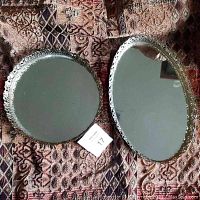 Oval and round vintage mirrored trays placed on patterned fabric, showing reflective surfaces and decorative edging.