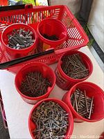 Multiple red containers containing assorted galvanized screws, bolts, nails, nuts, and washers. Some rust present, items stored on a white surface.