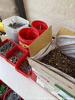 Photo showing an almost full cardboard box containing numerous 2 1/2 inch yellow zinc screws with surrounding red and white plastic containers holding various metal fasteners, staples, screws.