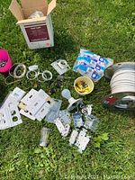 Photo showing partial rolls of white electrical wire, various electrical outlets, switch covers, light bulb, and packages of wire staples arranged outside on grass.