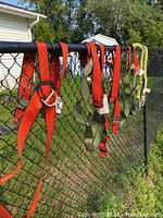 Three safety harnesses hanging on chain link fence: two red and one green. Visible metal D-rings and adjustable straps.
