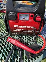 Front view of Motomaster Mobile Power Pack showing red front panel, analogue DC voltmeter, black handle, and battery clamps (red and black) connected to cables.
