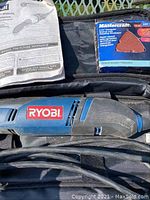 Blue and black Ryobi electric detail sander laying inside black cloth zipper case, with attached power cord visible. Accompanied by user manual and box of Mastercraft sanding sheets.