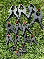 Full view of 14 assorted black plastic spring clamps with red tips arranged on grass showing varying sizes.