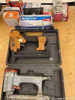 Two pneumatic nailers in black carrying cases on a workbench; one yellow Stanley Bostitch stapler showing wear and one silver Paslode nailer with some paint scuffs.