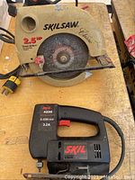 Photo of beige Skilsaw Classic circular saw showing blade and power cord with yellow plug, placed on wooden surface with black Skil jigsaw partially visible