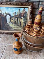 Photo showing the oil painting framed in brown wood, wooden bucket with metal bands, wooden vase with blue flowers, and a wooden drink set with jug and six cups decorated with red and black patterns.