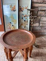 Folding wooden table with engraved round tray and metal candle holder in front of stone fireplace with two framed shell art pictures behind.