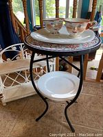 Full view of tile top metal table holding two ceramic face cups on a smiling platter and a plain white plate on lower shelf.