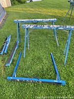 Overview of six blue metal folding sawhorses on grass, some folded, some standing, with paint stains.