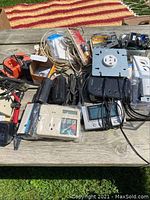 Wide view showing assorted electrical components on wooden table outside, including power strips, outlets, panel meter, and chargers.