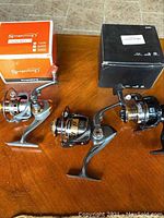 Three new fishing reels placed on a wooden surface. Two reels with folding handles, one with extended handle with large foam knob. Two original boxes are visible: an orange Sougayilang box and a black box.