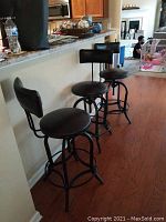 Three black bar stools lined up along a kitchen counter showing cushioned round seats, padded backrests, and metal frames with footrests.
